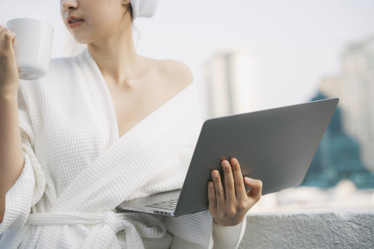 穿着白色浴袍的年轻女子早上在家拿着笔记本电脑喝热咖啡。在家工作。