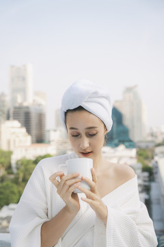 身着白色浴袍的年轻女子在城市背景的公寓阳台上享用热咖啡。
