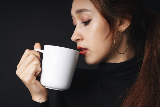 在黑暗的背景下，穿着黑色毛衣的漂亮年轻女子的肖像。拿着白色杯子。咖啡或茶。从杯子里喝水的女人。