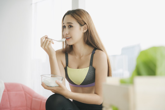 穿着运动装的漂亮女人，自然妆容，白色背景。穿着运动内衣的健康女性喜欢在沙发上用玻璃碗里的勺子吃酸奶。