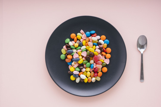 顶视图-黑色盘子上五颜六色的糖果，粉红色背景上有勺子。作为食物的甜点。