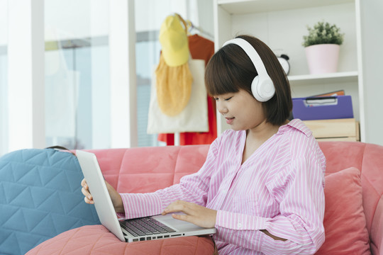 年轻的亚洲学生黑色短发女子戴着耳机在客厅的沙发上用笔记本电脑在家学习。