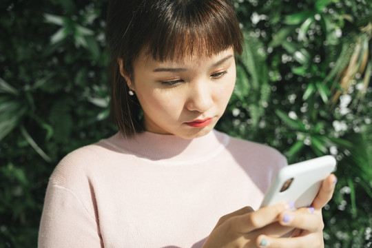 美丽的亚泰女人穿着粉色衬衫，在绿色自然背景下使用智能手机。