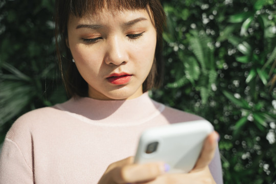 美丽的亚泰女人穿着粉色衬衫，在绿色自然背景下使用智能手机。