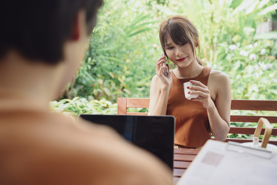 快乐的亚泰女人在户外打电话喝着茶，和正在笔记本电脑上工作的男朋友坐在一起。