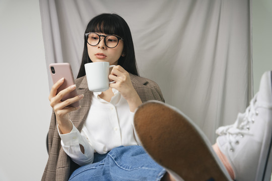 年轻漂亮的亚泰裔黑发女商人手里拿着一杯咖啡，用着智能手机。时尚肖像。