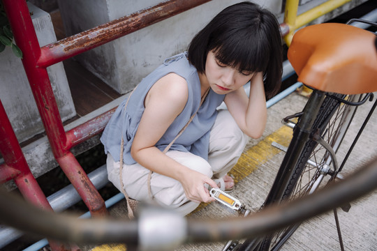 一位年轻的泰国亚裔女子坐在一辆坏了的自行车旁，沮丧地等着别人来修理。