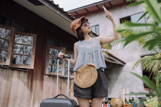 美丽的亚泰女旅行家，穿着夏季休闲服，戴着帽子，带着当地酒店的手推车行李。本地旅行。