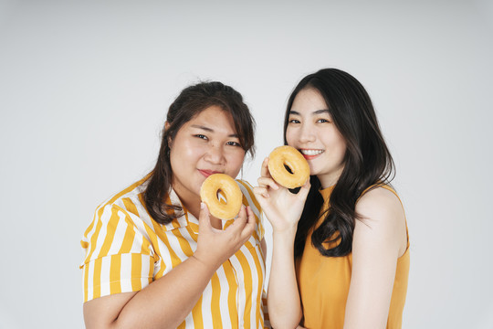 两个亚洲胖乎乎的苗条女人拿着不健康的甜甜圈，在白色背景下隔离开来。