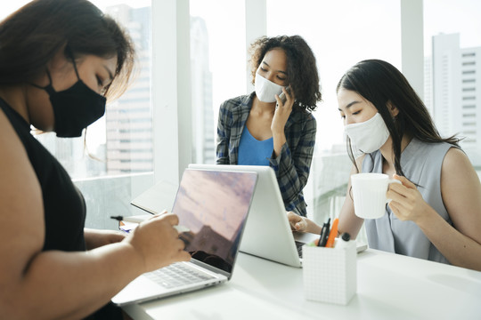 三位不同的亚非商界女性戴着面具一起在办公室工作。