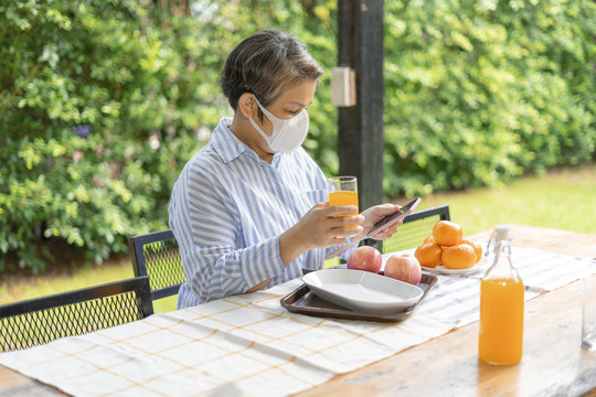 老年妇女戴着智能手机口罩，在院子里拿着橙汁杯。