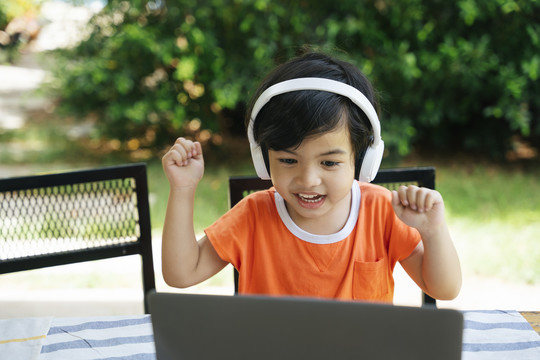兴奋的亚洲小孩在户外院子里戴着耳机用笔记本电脑。