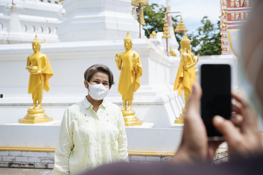 亚洲老年夫妇戴着口罩在泰国寺庙里用智能手机拍照。