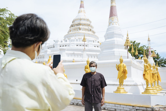 亚洲老年夫妇戴着口罩在泰国寺庙里用智能手机拍照。