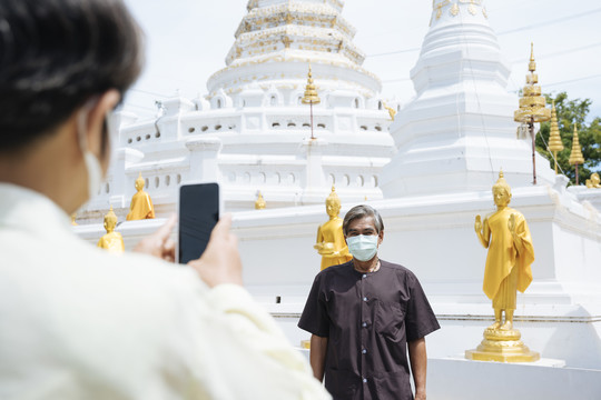 亚洲老年夫妇戴着口罩在泰国寺庙里用智能手机拍照。