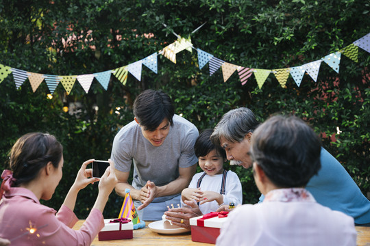 亚洲小孩在户外和家人一起庆祝生日聚会。妈妈用智能手机拍照。