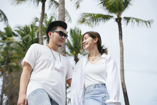 一对穿着白衬衫的亚洲情侣手牵着手在沙滩上散步。