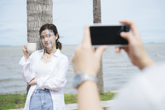 在海滩上的椰子树上，一名男子手持咖啡杯，戴着面罩，手持智能手机与美丽的亚洲女子合影。
