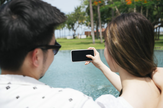 后视图-亚洲情侣旅行者在游泳池用智能手机自拍。
