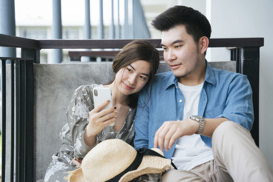 坐在酒店阳台上使用智能手机的亚洲情侣旅行者。