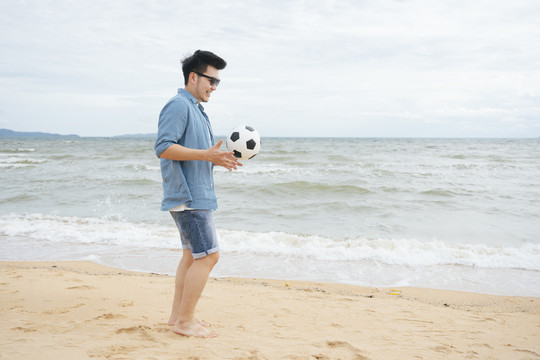 一个亚洲旅行者，穿着休闲衬衫，戴着墨镜，拿着足球在沙滩上散步。