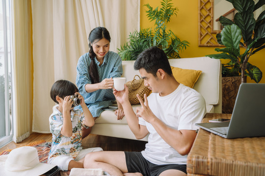 一个亚洲家庭在家共度时光。儿子练习用相机对着父亲拍照。妈妈给爸爸一杯水。