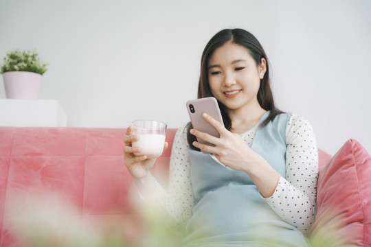 年轻漂亮的亚洲孕妇在客厅里用智能手机拿着一杯牛奶。