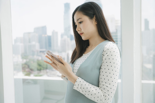 侧视图-年轻美丽的泰国亚裔孕妇使用智能手机查看社交媒体。