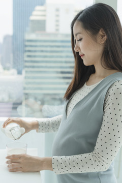 侧视图-年轻美丽的亚洲孕妇从瓶子里倒鲜奶。