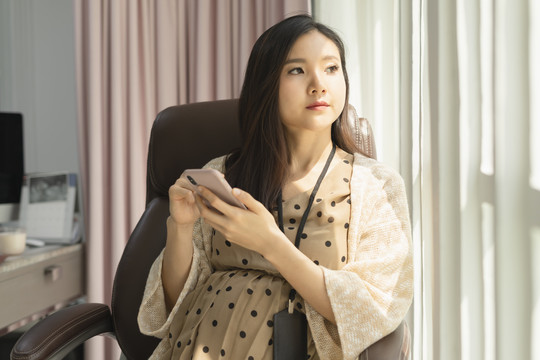 美丽的亚洲女商人坐在办公室的窗户边用智能手机。
