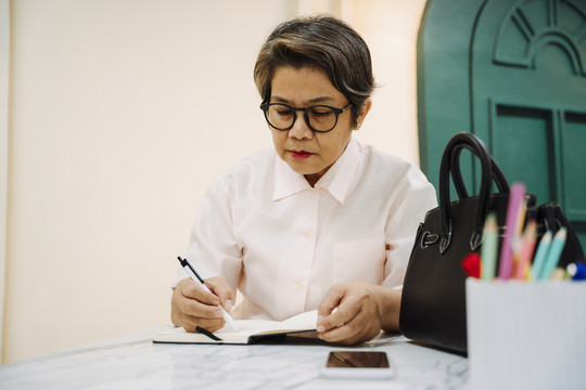 亚洲老年妇女戴眼镜在书上写日记。