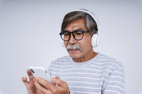 照片中的老人在白色背景下用无线耳机和智能手机听音乐。