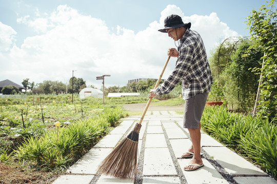 在一个农场里，一位亚洲老年农民用扫帚杆打扫花园。