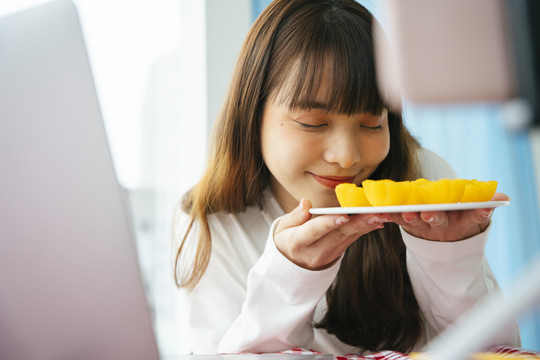 年轻的亚洲黑发女子在智能手机摄像头前拿着金色蛋糕做泰国甜点食品评论。