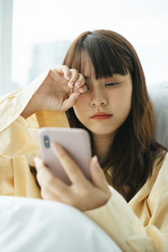 昏昏欲睡的亚洲黑发女青年早上醒来躺在床上用智能手机。