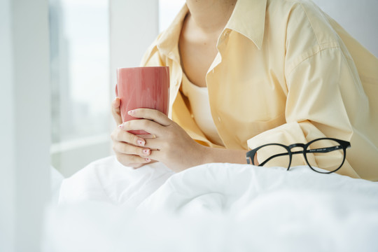 侧视图-年轻的亚洲女人早上在床上拿着一杯咖啡。