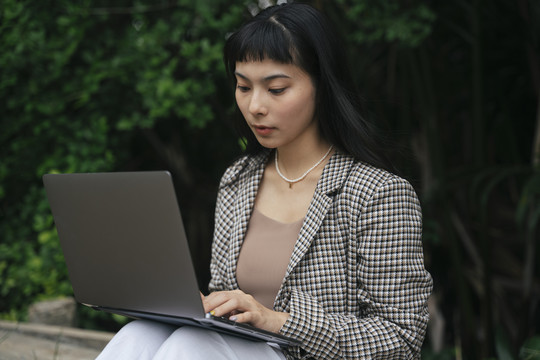 穿着正式衬衫的亚洲女商人在户外使用笔记本电脑。