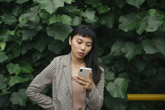 穿着正式衬衫的亚洲女商人在自然背景下使用智能手机。