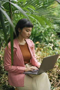 穿着正式衬衫的亚洲女商人坐在椅子上用笔记本电脑。