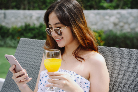 美丽的亚洲女人用智能手机戴着眼镜，在酒店的沙滩椅上拿着橙色的眼镜。