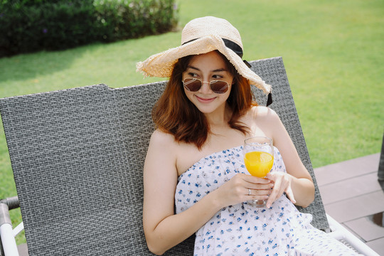 美丽的亚洲女人戴着帽子和太阳镜在沙滩椅上放着橙汁放松。