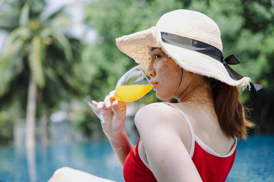 侧视图-美丽的亚洲女游客穿着红色泳衣和帽子在游泳池喝橙汁。