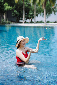 美丽的亚洲女游客穿着红色泳衣，戴着帽子在游泳池里泼水。