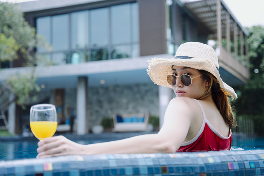美丽的亚洲女游客戴着墨镜和帽子，在游泳池里喝着一杯橙汁放松。