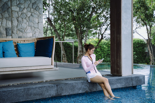 亚洲女游客穿着游泳衣在度假村和酒店的泳池边打电话。