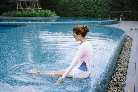 美丽的亚洲女游客穿着游泳衣坐在室外游泳池边。