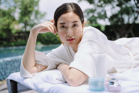 在游泳池旁的水疗床上，一位穿着白色浴袍的亚洲妇女的肖像，她拿着化妆瓶。