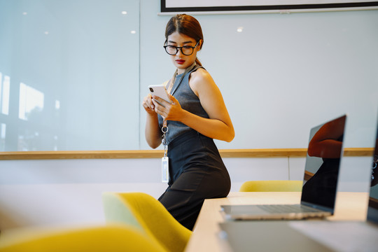 戴眼镜的亚洲女商务人士用手机，坐在会议室的桌子上。