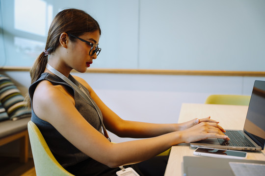 侧视图-亚洲女商人戴眼镜在笔记本电脑上打字。