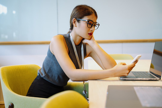 侧视图-亚洲女商人在办公室使用智能手机戴眼镜。